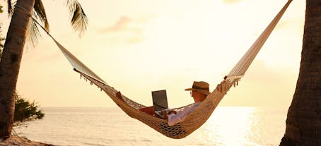 Woman business traveler lying in hammock on the tropical beach at sunset, working remotely during summer vacation.
