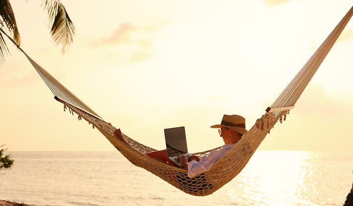 Woman business traveler lying in hammock on the tropical beach at sunset, working remotely during summer vacation.